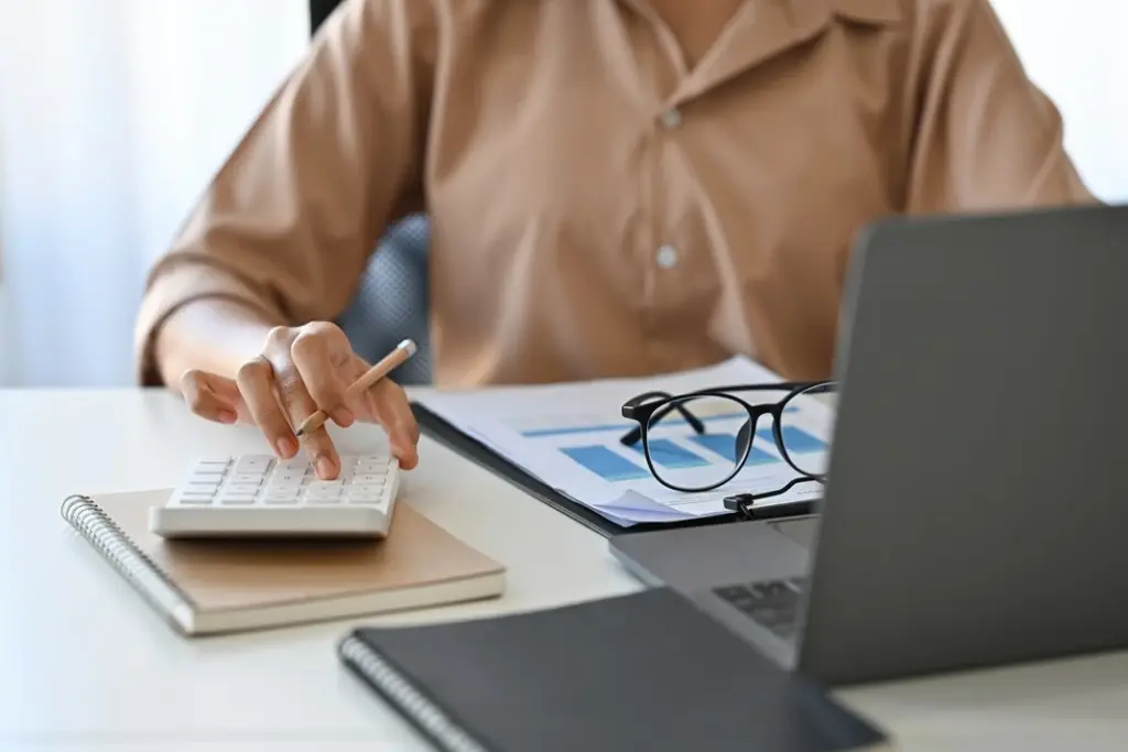 Close-up of a person using a calculator while reviewing financial documents and working on a laptop, representing accounting tasks.