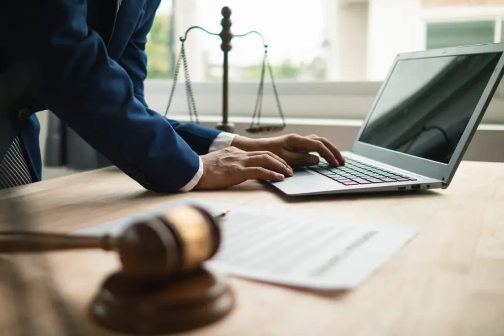 Close-up of a legal professional's hand working on a laptop, with a gavel and scales of justice in the background.