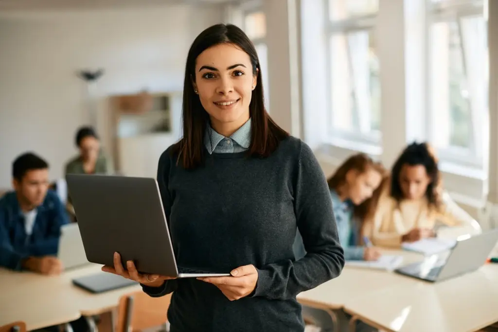 Smiling educator holding a laptop in a classroom, with students engaged in learning in the background.